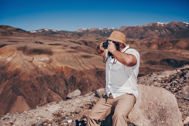 Fotografiando montañas rocosas. Un hombre adulto está sentado en una roca con una cámara digital en sus manos y filmando el gran cañón bajo un cielo claro y sin nubes.