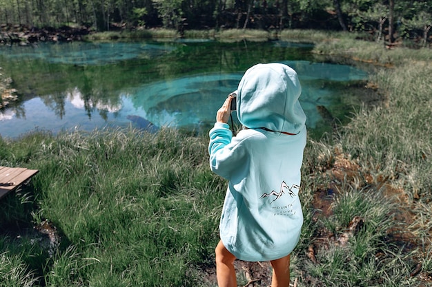 Fotografiando la naturaleza del bosque en un viaje. Una joven con capucha azul y una capucha está parada con la espalda y sosteniendo una cámara en sus manos. Un paisaje de verano con un estanque claro, árboles y césped.