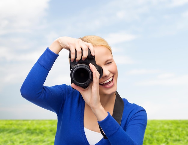 Fotografie-, Technologie-, Sommer- und Personenkonzept - lächelnde junge Frau, die Foto mit Digitalkamera über blauem Himmel und Grashintergrund macht