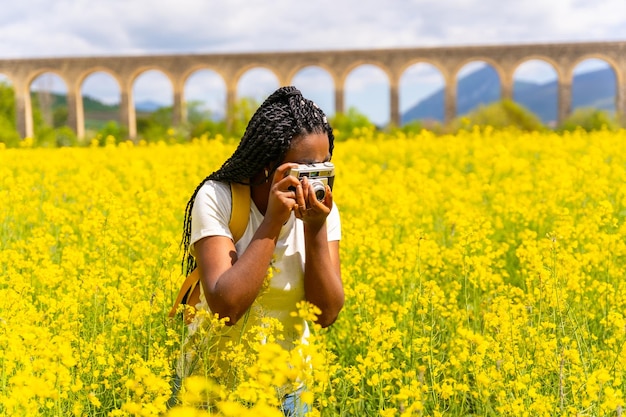 Fotografieren mit einer Vintage-Kamera ein schwarzes ethnisches Mädchen mit Zöpfen ein Reisender in einem Feld mit gelben Blumen