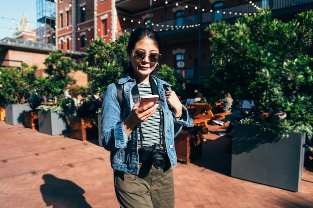 Fotografin eines jungen Mädchens, das ein Handy hält, das Richtung Online-Karte zum Hotel sucht und an einem sonnigen Tag auf rotem Backsteinboden in der geschäftigen Stadt San Francisco spazieren geht. schöne asiatische Frau mit Sonnenbrille lächelnd.