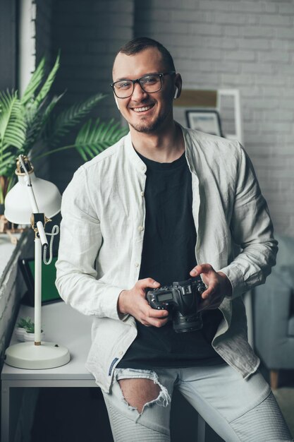 Fotógrafo alegre segurando a câmera sorrindo para a câmera em pé em seu escritório Trabalho criativo freelance marketing on-line Inicialização Proprietário de pequenas empresas