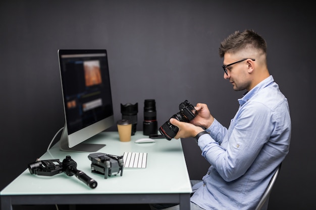 Fotógrafo confiado. Joven alegre sosteniendo una cámara digital y sonriendo mientras está sentado en su lugar de trabajo