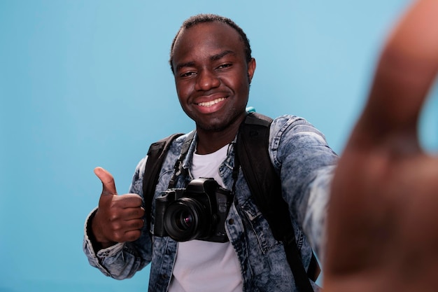 Fotógrafo confiado que muestra el símbolo del dedo bien mientras toma selfie y tiene una cámara DSLR en fondo azul. Entusiasta de la fotografía que aprueba el gesto de la mano mientras se toma una foto.