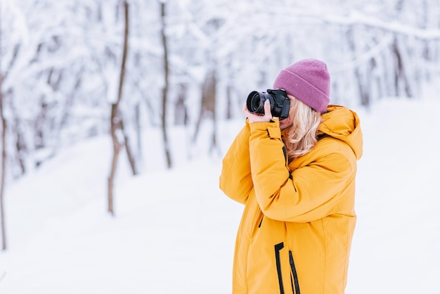 Fotógrafo de garota feliz em uma jaqueta amarela tira fotos do inverno em um parque nevado
