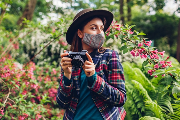 Fotógrafo de mulher tirando fotos na câmera digital no jardim de verão usando máscara. Freelancer gosta de filmar plantas florescendo