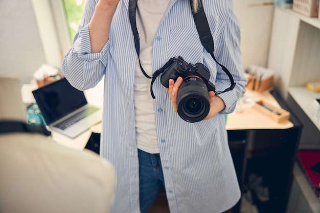 Fotógrafo experto en camisa sosteniendo una cámara de alta calidad mientras está de pie en la sala de trabajo
