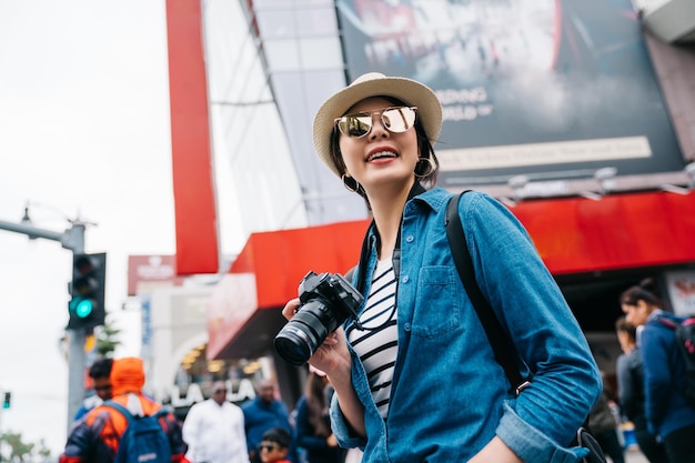 fotógrafo feminino atraente em pé na rua movimentada e tirando foto dos moradores. linda jovem segurando a câmera dslr. estilo de vida de mulher de lente de viagem.