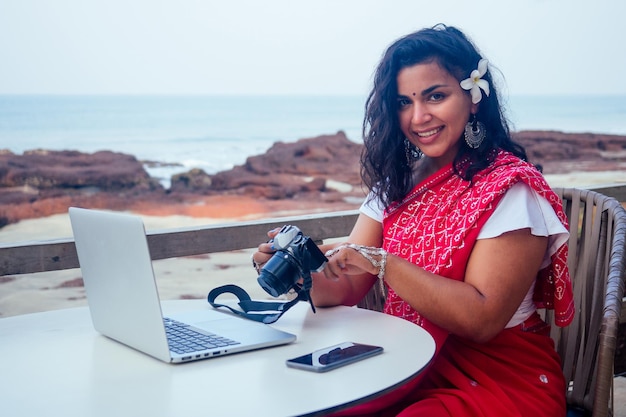 Fotógrafo feminino da Índia fotografando com câmera digital na praia mulher de negócios mulher indiana em sari tradicional vermelho no mar do paraíso tropical tirando fotos da sessão de fotos da paisagem