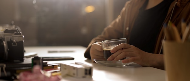 Fotógrafo feminino segurando a xícara de café enquanto está sentado na mesa de trabalho com câmera e suprimentos em estúdio