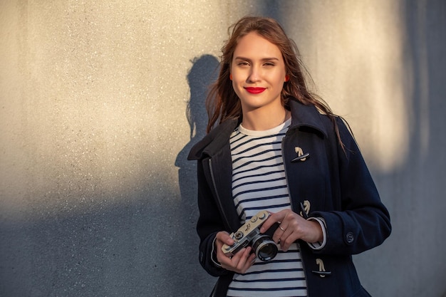 Fotógrafo feminino sorridente na jaqueta em frente à parede pronta para fazer uma nova foto. Adorável jovem morena com roupa da moda posando no fundo da parede de concreto com câmera