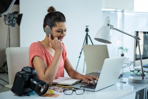 Fotógrafo feminino trabalhando sobre o laptop enquanto fala no celular