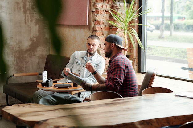 Fotógrafo inconformista con gorra de béisbol sentado a la mesa en el café y discutiendo el plan de sesión de fotos con el asistente después de la cuarentena por coronavirus