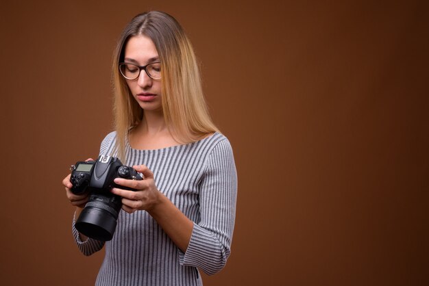 Fotógrafo joven y bella mujer sosteniendo una cámara DSLR contra la pared marrón