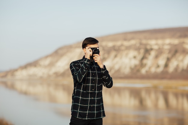 Fotógrafo joven elegante tomando fotografías con cámara vintage en una montaña durante el viaje.