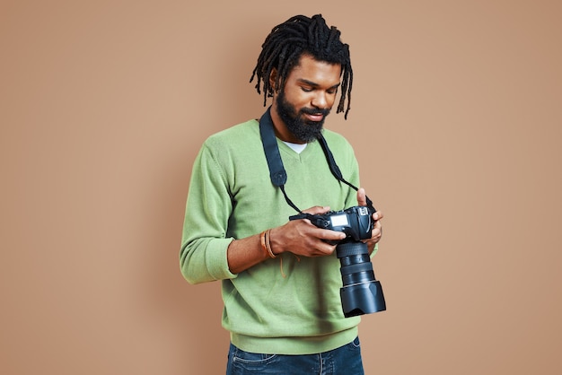 Fotógrafo joven guapo en ropa casual con cámara digital y sonriendo mientras está de pie contra la pared marrón