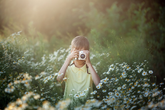 El fotógrafo lindo del muchacho tira en cámara en naturaleza. Kid toma una foto en el campo de flores de manzanilla.