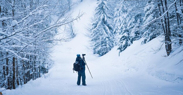 Un fotógrafo masculino ajusta su cámara en un trípode para fotografiar el borde de un bosque de montaña en los hermosos Cárpatos en clima nevado