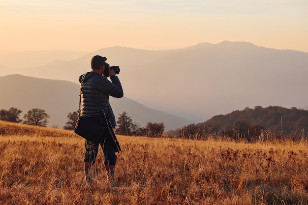 Fotógrafo masculino de pé e trabalhando na majestosa paisagem de árvores de outono e montanhas no horizonte