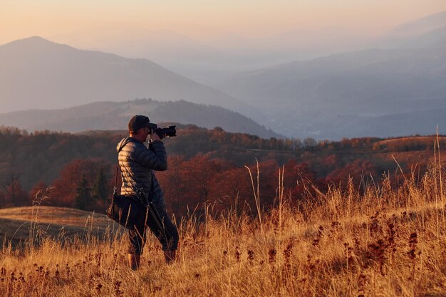 Fotógrafo masculino de pé e trabalhando na majestosa paisagem de árvores de outono e montanhas no horizonte