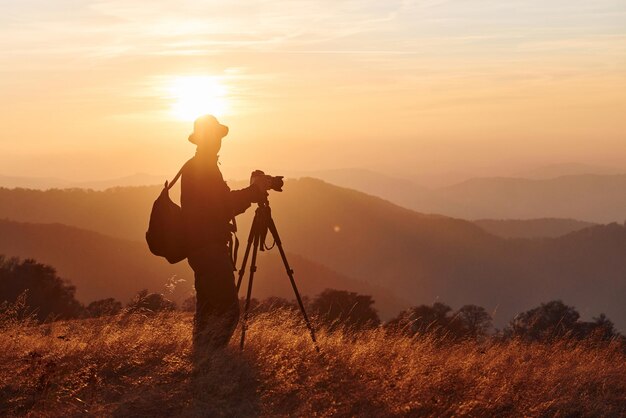 Fotógrafo masculino de pé e trabalhando na majestosa paisagem de árvores de outono e montanhas no horizonte