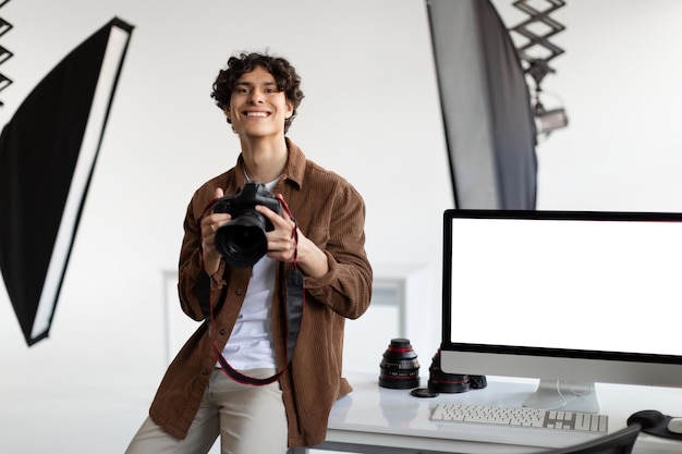 Fotógrafo masculino feliz con cámara profesional sentado en su escritorio con monitor de computadora con maqueta de pantalla en blanco