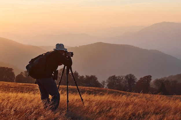 Fotógrafo masculino de pie y trabajando en el majestuoso paisaje de árboles y montañas de otoño en el horizonte