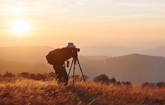 Fotógrafo masculino de pie y trabajando en el majestuoso paisaje de árboles y montañas de otoño en el horizonte