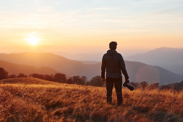 Fotógrafo masculino de pie y trabajando en el majestuoso paisaje de árboles y montañas de otoño en el horizonte