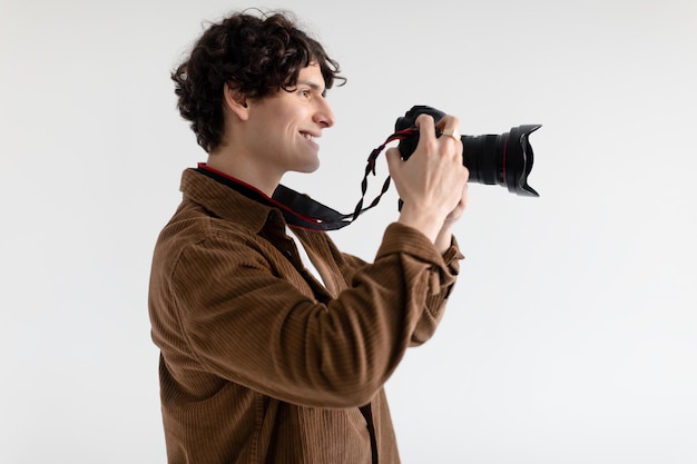 Fotógrafo masculino positivo tirando foto segurando a câmera perto do rosto e sorrindo em pé sobre o branco