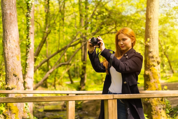 Fotógrafo de mujer con una chaqueta negra disfrutando en un parque de otoño, tomando fotografías al atardecer en un puente de madera