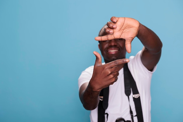 Fotógrafo profesional confiado haciendo gestos de marco con las manos mientras tiene una cámara moderna sobre fondo azul. Chico joven con dispositivo DSLR gesticulando tomando fotos con las manos. foto de estudio