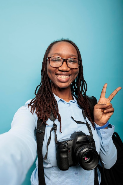 Fotógrafo sonriente feliz con un dispositivo de cámara moderno que da un signo de paz mientras toma selfie. Entusiasta de la fotografía grabando video vlog diario mientras está de pie sobre fondo azul. foto de estudio