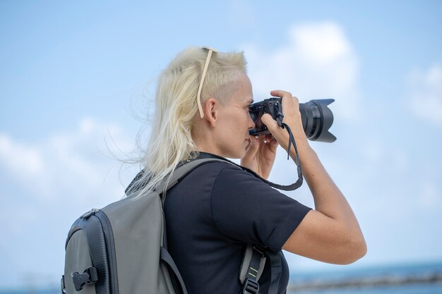 Fotógrafo de viajero turístico tomando fotografías del paisaje en la cámara de fotos, chica hipster con mochila disfrutando del hermoso paisaje del mar. Cerrar retrato joven rubia con mochila en la playa