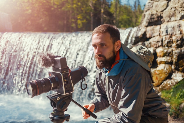 Foto fotógrafo de viajes hombre barbudo de cerca con cámara de película profesional en trípode disparando paisaje de montaña en el fondo de la cascada