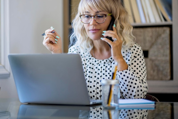 Fotografou uma empresária feliz sentada à mesa atrás de seu laptop e conversando com alguém em seu celular enquanto trabalhava em casa Escritório em casa Empresária serena falando ao telefone na mesa