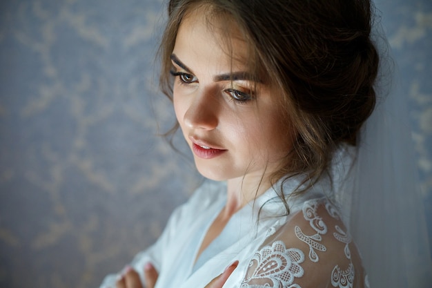 Fotonahaufnahmeporträt einer Braut mit professionellem Make-up, sie ist in einem weißen Mantel, Schleier. Hochzeitstag der jungen Frau. Fotoshooting, Frisur, Beauty