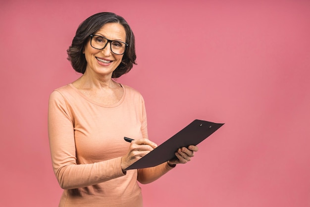 Fotoporträt der älteren reifen gealterten Geschäftsfrau konzentrierte sich, die Zwischenablage und das Lesen des Stiftes lokalisiert über rosafarbenem Hintergrundexemplar hielt.