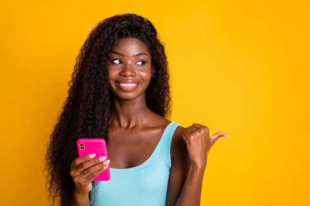 Fotoporträt einer aufgeregten afroamerikanischen lockigen brünetten Frau, die mit dem Daumen auf eine Leerstelle zeigt, die rosa Telefon in der Hand hält, isoliert auf leuchtend gelbem Hintergrund