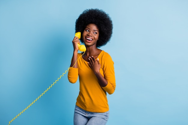 Fotoporträt einer schwarzhäutigen Frau, die am Retro-Telefon spricht und den gelben Hörer mit Draht isoliert auf leuchtend blauem Hintergrund hält