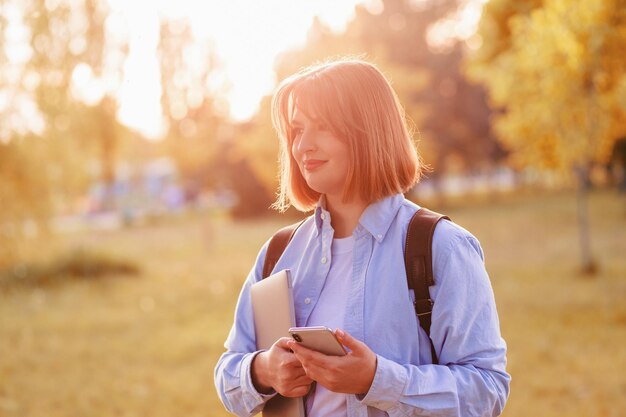 Fotoporträt einer Studentin mit Telefon und Laptop, die draußen im Park lächelt