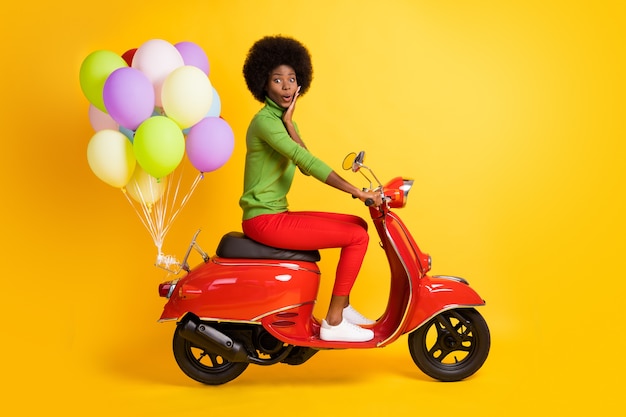 Fotoporträt eines beeindruckten brünetten dunkelhäutigen Mädchens mit grünem Pullover auf rotem Fahrrad mit bunten Luftballons einzeln auf leuchtend gelbem Hintergrund