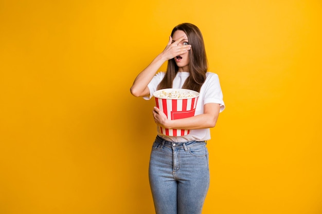 Fotoporträt eines verängstigten Mädchens, das Popcorn-Karton hält und Horror mit geschlossenem Gesicht beobachtet, mit hand isoliertem leuchtend gelbem Farbhintergrund