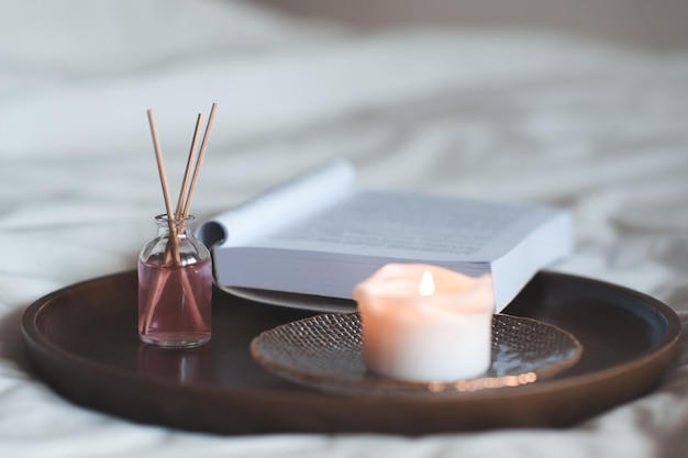 Fragancia líquida casera en botella de vidrio con palos de madera y velas aromáticas con libro de papel abierto en la bandeja en la cama de cerca