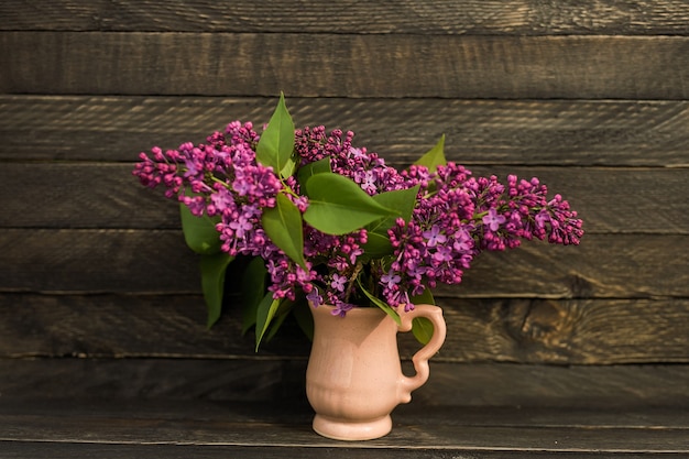 Un fragante ramo de lilas se encuentra en una pequeña jarra rosa en una pared de madera en un estilo rústico.