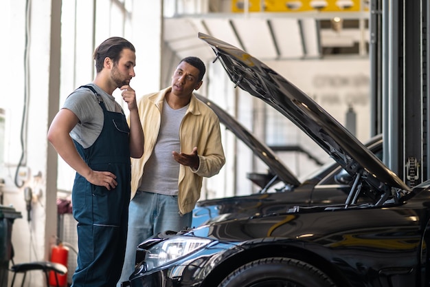 Frage. Nachdenklicher kaukasischer Mechaniker, der das Kinn berührt und in die Motorhaube schaut, und dunkelhäutiger Kunde, der in der Nähe des Autos steht und fragt