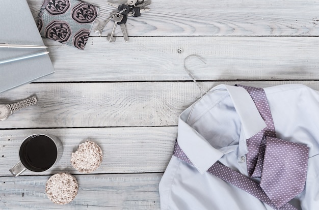 Fragmento de una camisa de hombre con corbata en una percha, galletas, taza de café sobre una superficie de madera pintada. Los colores pastel