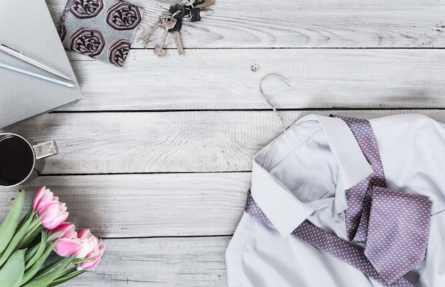 Fragmento de una camisa de hombre con corbata en una percha, tulipanes, taza de café sobre una superficie de madera pintada. Los colores pastel