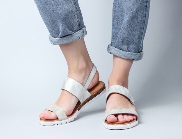 Fragmento de piernas femeninas en jeans y sandalias de cuero de moda en blanco. Zapatos de verano con estilo para mujer.