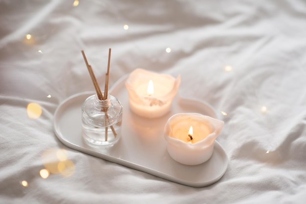 Fragrância líquida doméstica em garrafa de vidro e velas acesas que ficam na bandeja de cerâmica branca na cama fecham a atmosfera aconchegante e hygge
